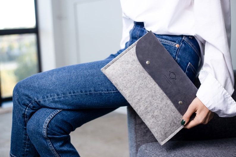 May include: A gray felt laptop sleeve with a black leather flap and a silver metal button. The sleeve has a logo that says "Simple & Sustainable".