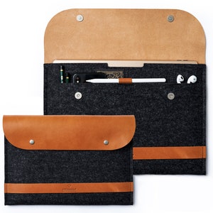 Può includere: Due custodie per tablet in feltro grigio con dettagli in pelle marrone. Una custodia è aperta, rivelando un tablet, una penna, un telefono e auricolari. L'altra custodia è chiusa. Le custodie hanno chiusure magnetiche e un cinturino in pelle marrone.