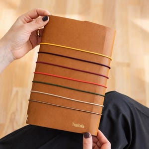 Könnte beinhalten: Ein braunes Notizbuch mit bunten Gummibändern. Das Notizbuch wird von einer Person gehalten. Der Text "Tusitala" ist unten rechts auf dem Notizbuch aufgedruckt. Die Gummibänder sind gelb, lila, rot, grün und beige.