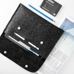 May include: A gray felt laptop sleeve with a zipper closure and a black leather bottom. The sleeve is open and a tablet, a stylus, and a blue pencil are inside. A newspaper and a black card with the word "Oyster" are on the white surface.