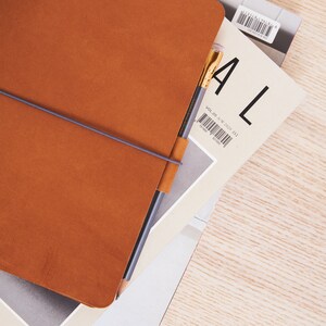 May include: A brown leather bound journal with a grey elastic band and a pencil sticking out of the top. The journal is on top of a magazine with the word "ALL" printed on the cover.
