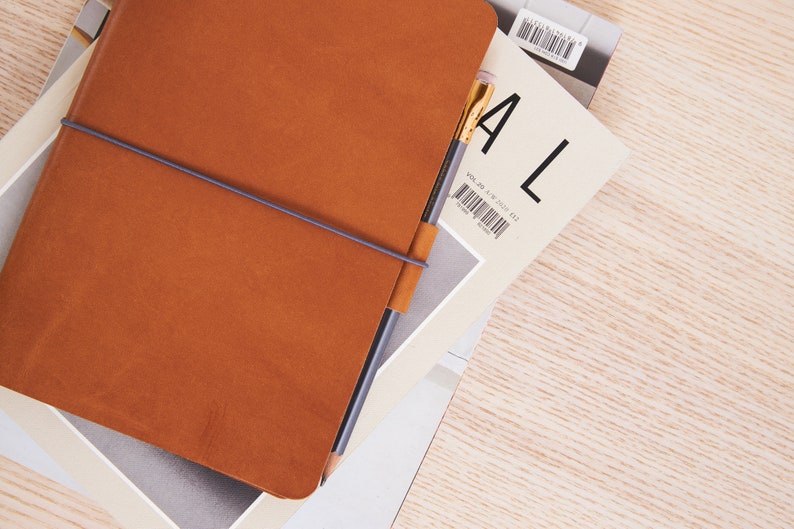 May include: A brown leather bound journal with a grey elastic band and a pencil sticking out of the top. The journal is on top of a magazine with the word "ALL" printed on the cover.