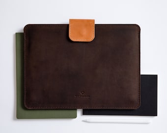 May include: A brown leather laptop sleeve with a tan leather flap closure. The sleeve has a stitched logo that reads "CITYSHEEP" in white text. The sleeve is sitting on top of a green notebook and a black notebook.