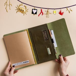 Op de afbeelding: Een open olijfgroen leren notitieboek met een pen en papier erin. Boven het notitieboek hangt een slinger van goudkleurige bedels, waaronder een krab, octopus en hart. Het notitieboek wordt door twee handen opengehouden.