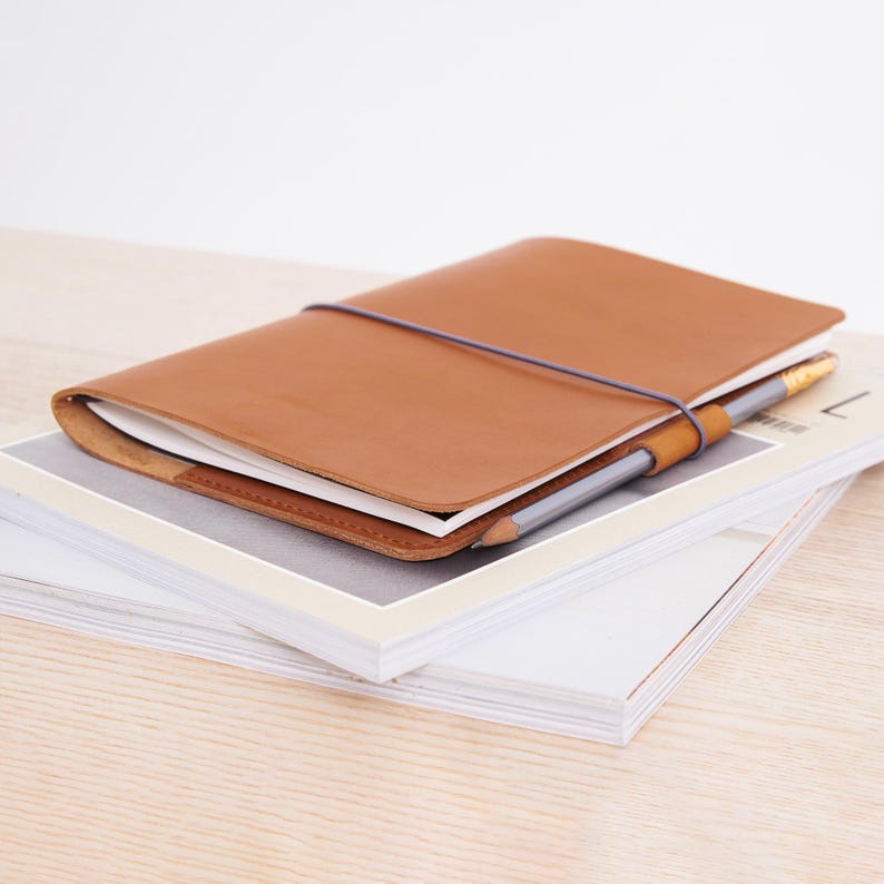 May include: A brown leather notebook with a pencil and elastic band. The notebook is on top of a stack of white papers.