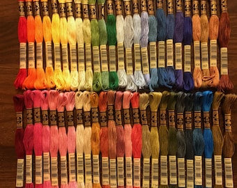 Puede incluir: Una colección de carretes de hilo de bordar DMC en varios colores, dispuestos en filas. Cada carrete tiene una etiqueta con un número de color DMC. Los colores van desde el amarillo brillante hasta el azul oscuro.