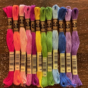May include: A collection of 12 DMC embroidery floss spools in various colors, including pink, orange, yellow, green, blue, and purple. Each spool has a white label with black text, including the DMC number and the brand name.