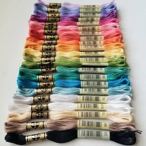 May include: A collection of 20 spools of DMC embroidery floss in various colors, including pink, blue, green, yellow, and beige. Each spool has a black and gold label with the DMC number printed on it.