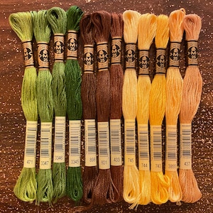 May include: A collection of 12 DMC embroidery floss spools in various shades of green, brown, yellow, and orange. Each spool has a white label with black text, including the DMC number, the color name, and the length of the floss. The spools are arranged in a row on a wooden surface.