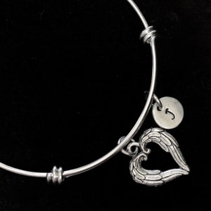 Può includere: Bracciale regolabile color argento con un ciondolo a forma di cuore con dettagli alati. Un piccolo ciondolo rotondo con la lettera "J" è appeso vicino. Il bracciale è posto su uno sfondo nero.