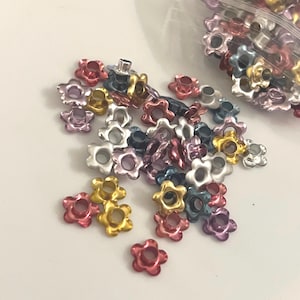 May include: A collection of colorful flower-shaped metal rivets in various shades, including pink, gold, silver, and blue. The rivets are scattered on a white surface, with some contained in a clear plastic bag. These decorative rivets are ideal for craft projects.