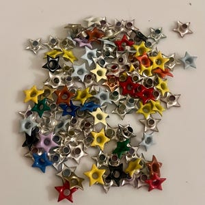 May include: A collection of star-shaped metal rivets in various colors, including red, yellow, green, blue, and black. The rivets have a silver-colored metal base and are scattered on a white surface. These decorative rivets are ideal for crafts and DIY projects.