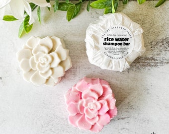 Puede incluir: Dos barras de champú en forma de flor, una blanca y otra rosa, con el texto "rice water shampoo bar" en una etiqueta blanca. La etiqueta también dice "sulfate-free unscented" y "HANDMADEINCA.COM".