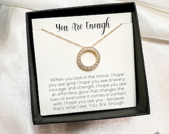 May include: A gold necklace with a circle pendant encrusted with clear rhinestones. The necklace comes in a small box with a white card that reads "You Are Enough" and a message about seeing your own strength and beauty.