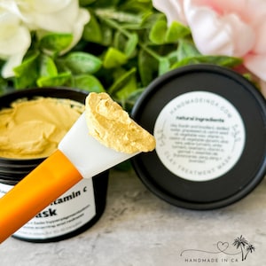 May include: A close-up of a jar of yellow face mask with a white spatula scooping out a dollop of the mask. The jar is labeled "Vitamin C Mask" and the lid of the jar says "Handmadeinca.com Natural Ingredients Clay Treatment Mask".