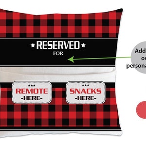 May include: Red and black buffalo plaid pillow with a white pocket and the text "RESERVED FOR" with an arrow pointing to the right. The pocket has two white labels with the text "REMOTE -HERE- " and "SNACKS -HERE-"
