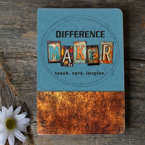 May include: A blue notebook with a rusty metal design. The notebook has the words "Difference Maker" in a circle with the word "Maker" spelled out in individual letters. The text "teach. care. inspire." is below the circle.