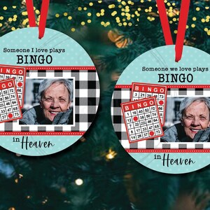 以下が含まれることがあります： 黒と白のバッファローチェック柄の丸い青と白のクリスマスオーナメント。オーナメントには女性の画像と2つのビンゴカードがあり、「愛する人が天国でビンゴをしている」というテキストが書かれています。