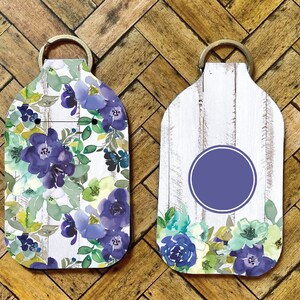 May include: Two keychains with floral designs. One features a pattern of purple and blue flowers with green leaves on a white wood-grain background. The other has a purple circle on a white wood-grain background with floral accents.