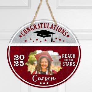 Peut inclure: Une suspension de porte ronde rouge et blanche avec le texte "Félicitations" et un bonnet de diplômé. Le texte "Reach for the Stars" est également sur la suspension. Une photo d'un diplômé est au centre de la suspension avec le texte "2025 Carson" en dessous de la photo.