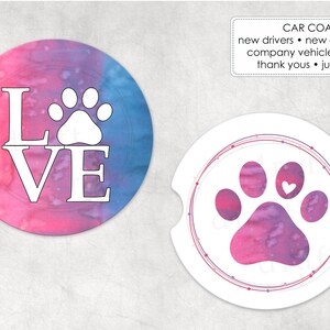 May include: Two round car coasters with a watercolour design. One coaster features a white paw print and the word "LOVE" in white. The other coaster features a pink and purple paw print with a white heart in the centre.
