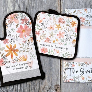 Könnte beinhalten: Ein Set aus drei Küchenartikeln mit einem Blumenmuster. Das Set beinhaltet einen Topflappen, einen Ofenhandschuh und ein Geschirrtuch. Der Topflappen und der Ofenhandschuh haben den Text "Happiness is... homemade." Das Geschirrtuch hat den Text "The Smiths" und ein Blumenmuster.