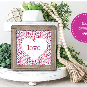 May include: A white wooden tiered tray with a framed print that says "love" in a pink heart shape on a background of colorful confetti. The tray is decorated with greenery, a wooden bead garland, and a jute tassel.