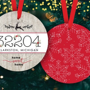 May include: Two round Christmas baubles. One bauble is white with a wood grain texture and features a black silhouette of a deer with red berries and the text "32204 CLARKSTON, MICHIGAN home sweet home". The other bauble is red with a white snowflake pattern.
