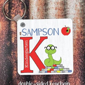 Könnte beinhalten: Ein weißer Holzschlüsselanhänger mit einem roten Buchstaben K, einer grünen Schlange mit Brille und einem roten Apfel mit einem Wurm. Der Text "MR. SAMPSON Kindergarten" steht auf dem Schlüsselanhänger.