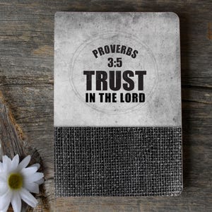 Puede incluir: Un diario gris y negro con una cubierta de arpillera. La portada presenta un círculo con el texto "Proverbios 3:5 Confía en el Señor".