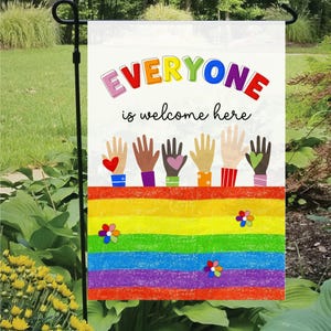 Peut inclure: Un drapeau de jardin avec le texte "EVERYONE is welcome here" aux couleurs de l'arc-en-ciel. Le drapeau présente un motif arc-en-ciel avec des mains et des fleurs colorées. Il est affiché à l'extérieur, dans un jardin.