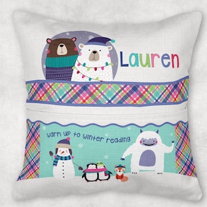 May include: A white pillow with a blue, green, and pink plaid pattern. The pillow has a pocket on the front and features a winter-themed design with a snowman, penguins, a yeti, and a fox. The text "Warm up to winter reading" is printed on the pillow. The name "Lauren" is printed on the pillow.