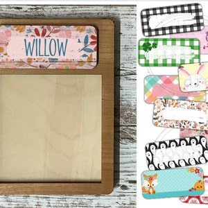 May include: A wooden frame with a blank white space inside. The frame has a pink and floral name tag on top that says "WILLOW". The image also shows 10 different designs for name tags, including owls, bunnies, ghosts, and foxes.