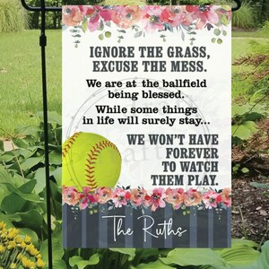 Może przedstawiać: Flaga ogrodowa z kwiatowym wzorem i piłką do softballu, z tekstem "IGNORE THE GRASS, EXCUSE THE MESS. We are at the ballfield being blessed. WE WON'T HAVE FOREVER TO WATCH THEM PLAY." Zawiera również imię "The Ruths."