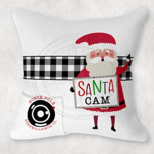 May include: White square pillow with a Santa Claus graphic holding a sign that reads "SANTA CAM." The pillow has a black and white buffalo plaid stripe and a North Pole Surveillance Co. logo.