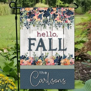 May include: A blue and white garden flag with a floral design and the text "hello, FALL" in a circle. The bottom of the flag has a blue background with white polka dots and the text "the Carlsons" in white cursive.