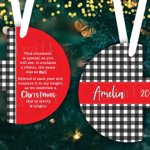 Puede incluir: Adorno navideño de búfalo a cuadros rojo y negro con una cinta blanca. El adorno tiene un mensaje personalizado que dice: "Este adorno es especial, como puedes ver. Contiene una cinta, del mismo tamaño que yo. ¡Desenvuélvelo cada año y compáralo con mi altura, mientras celebramos una Navidad alegre y brillante!" y "Amelia 2022".