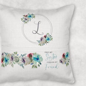Op de afbeelding: Een wit kussen met een zakje aan de voorkant. Het zakje heeft een bloemenpatroon met de tekst "First my Sister Forever my Friend". Het kussen heeft ook een bloemenpatroon met een cirkel en de letter "L" in het midden.
