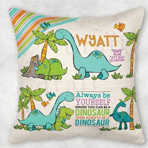 May include: A light beige cushion with a dinosaur theme. The cushion features a green and blue dinosaur with the text "Always be yourself unless you can be a dinosaur then always be a dinosaur". The cushion also features the name "Wyatt" and the text "Rawr means let's read in dinosaur."