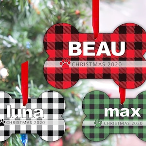 Pode incluir: Enfeites de Natal em forma de osso com padrões xadrez em vermelho, preto e verde. Cada enfeite tem o nome de um animal de estimação e "Christmas 2020" impresso. Uma fita vermelha é anexada para pendurar.
