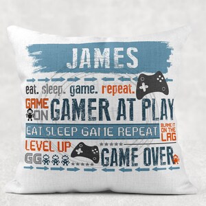 May include: White cushion with blue and orange text that reads "JAMES eat. sleep. game. repeat. GAME ON GAMER AT PLAY EAT SLEEP GAME REPEAT. LEVEL UP GG GAME OVER" with a blue and black video game controller graphic.