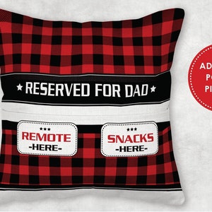May include: Red and black plaid pillow with a white pocket and the text "RESERVED FOR DAD" and two white labels that say "REMOTE -HERE- " and "SNACKS -HERE-"