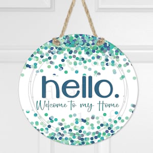 May include: Round welcome sign with a white background and blue and green confetti pattern. The sign says "hello. Welcome to my home".