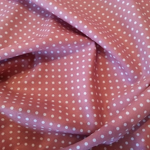 May include: A close-up shot of a dusty pink fabric featuring a repeating pattern of white polka dots. The fabric has a slightly wrinkled texture, creating shadows. The polka dots are evenly spaced and vary slightly in size.