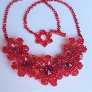 Op de afbeelding: Een rode kralenketting met een bloemenpatroon. De ketting is gemaakt van kleine, rode kralen die aan elkaar geregen zijn om een ​​reeks bloemen te vormen. De bloemen zijn in een cascade-patroon gerangschikt, met de grootste bloem onderaan en de kleinste bloem bovenaan. De ketting is afgewerkt met een eenvoudige sluiting.