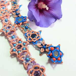 May include: Two beaded necklaces with star and flower designs. The necklaces are made with pink, blue and white beads. The necklaces are lying on a white surface next to a purple flower.