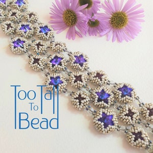 May include: A beaded bracelet with a floral design, featuring purple and white beads with a central purple crystal. The bracelet is displayed with purple flowers and the text "Too Tall To Bead".