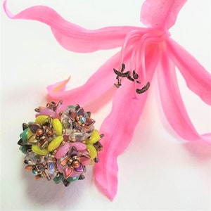 May include: A colourful beaded sphere with pink, yellow, green and brown beads, arranged in a flower-like pattern. The sphere is resting on a white surface next to a pink flower.