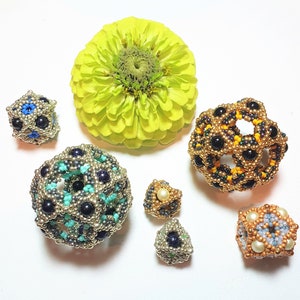 May include: Six beaded geometric shapes in various colors and sizes. The shapes are made of small beads in gold, black, blue, turquoise, and white. The shapes are arranged on a white surface with a green flower in the background.