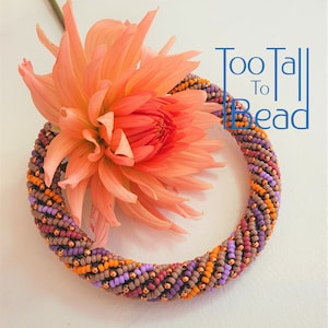 May include: A multi-coloured beaded bracelet with a spiral design. The bracelet is made with orange, brown, purple, and copper beads. The bracelet is lying on a white surface next to a peach-coloured flower.